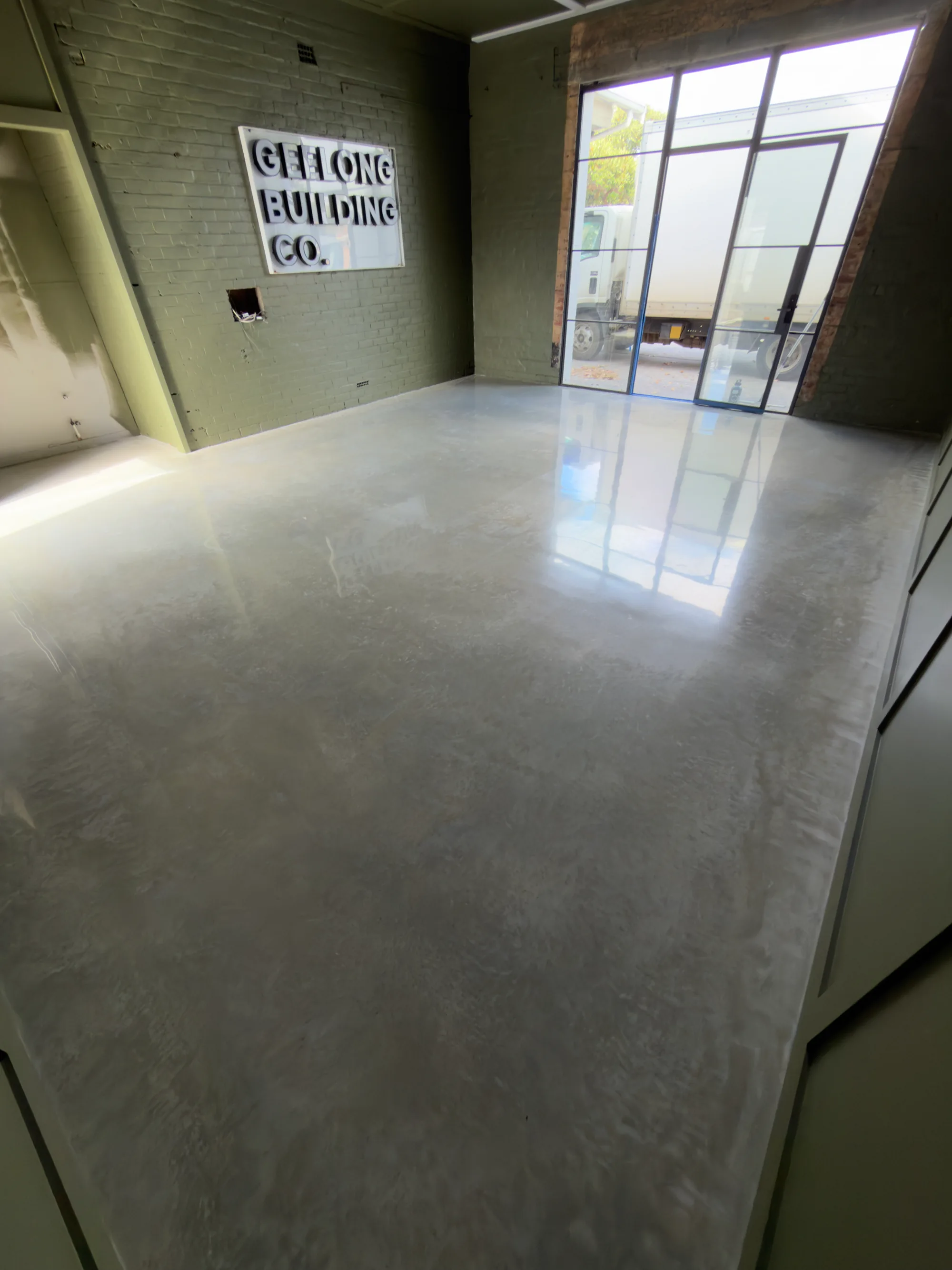 Surface-polished grey concrete floor reflecting a glazed doorway beneath the Geelong Building Co. wall sign